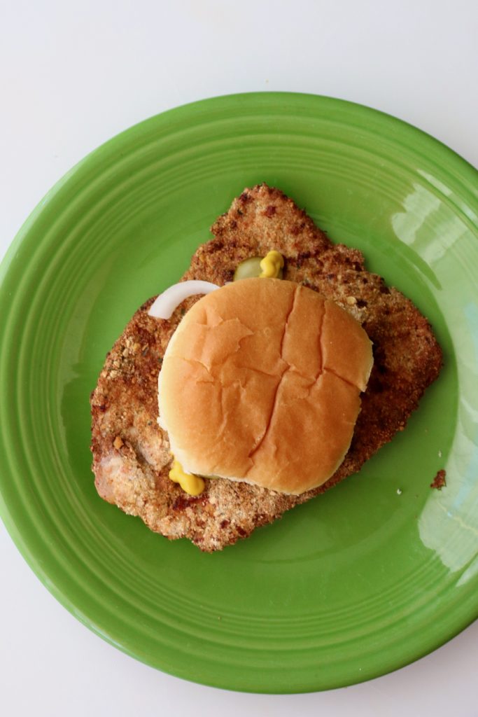 Pork tenderloin sandwich topped with mustard, onion and pickles.
