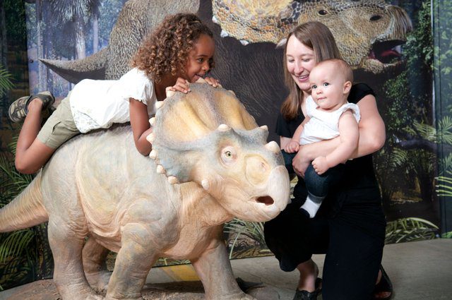 A mom holds a baby while a young girl lays on a dinosaur statue in the "Dinosaurs: Land of Fire and Ice" exhibit