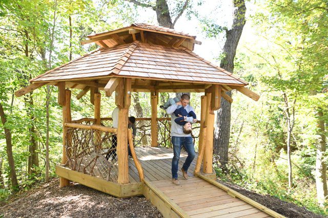 A dad holds a child upside down as they walk by a treehouse at the Treehouse Village at the Iowa Arboretum & Gardens